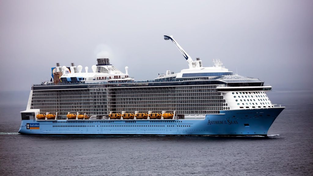 A wide side-profile shot of the Royal Caribbean cruise ship Anthem of the Seas sailing on calm open water under a misty sky, highlighting its light blue hull, rows of balcony cabins, and the signature North Star observation capsule arm on the top deck.