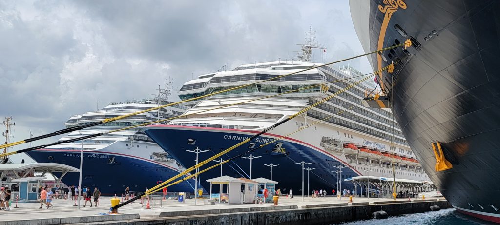 A close-up view of the bows of Carnival Conquest and Carnival Sunrise cruise ships docked side-by-side at a pier in Nassau, Bahamas under a cloudy sky.