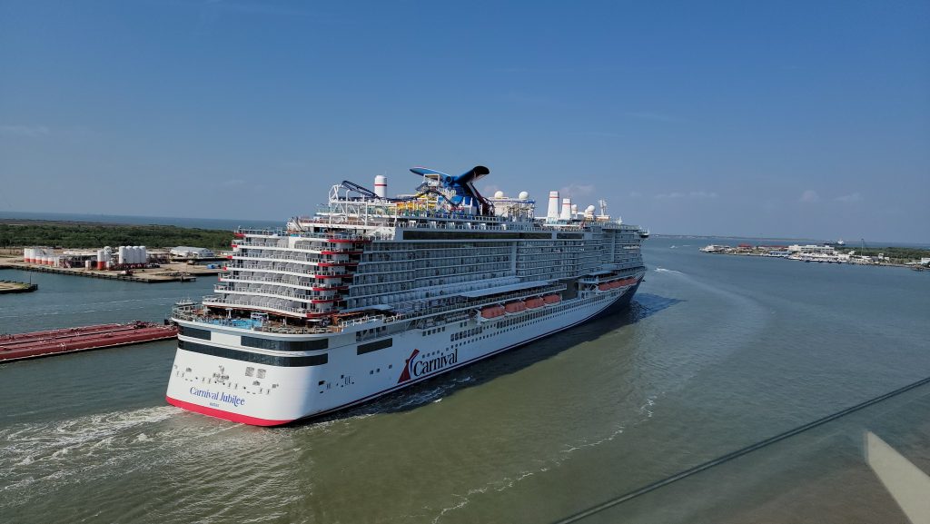 An elevated aft starboard view of Carnival Cruise Line's Excel-class ship, Carnival Jubilee, navigating a sunny shipping channel in Galveston, Texas, showcasing the top-deck BOLT roller coaster tracks and signature whale-tail funnel.