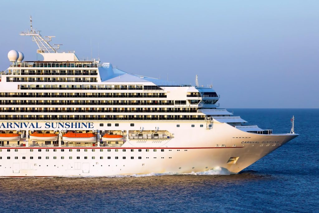 A close-up profile shot of the bow and forward decks of the Carnival Sunshine cruise ship sailing through calm, deep blue open ocean water during golden hour.