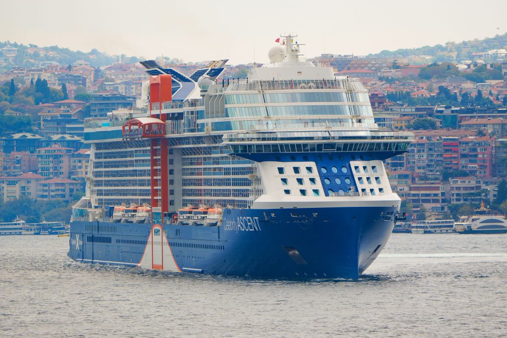 A large modern cruise ship, Celebrity Ascent, sailing through the Bosphorus Strait in Istanbul, Turkey, showcasing its dark blue and white hull, expansive forward glass windows, and signature orange Magic Carpet platform on the side with a hillside city in the background.