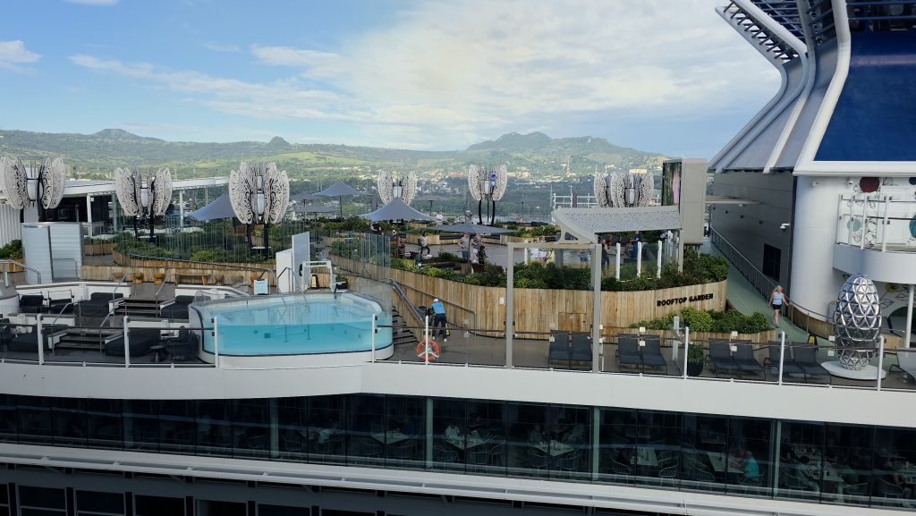 A close-up view of the lush greenery, seating areas, and unique metallic tree sculptures inside the Rooftop Garden on the Celebrity Beyond cruise ship, docked in Puerto Plata, Dominican Republic.