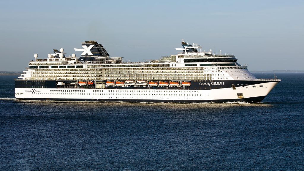 A side profile view of the Celebrity Cruises ship Celebrity Summit sailing on deep blue water under a clear sky, showcasing its white and dark blue hull and the iconic white 'X' on its dark funnel.