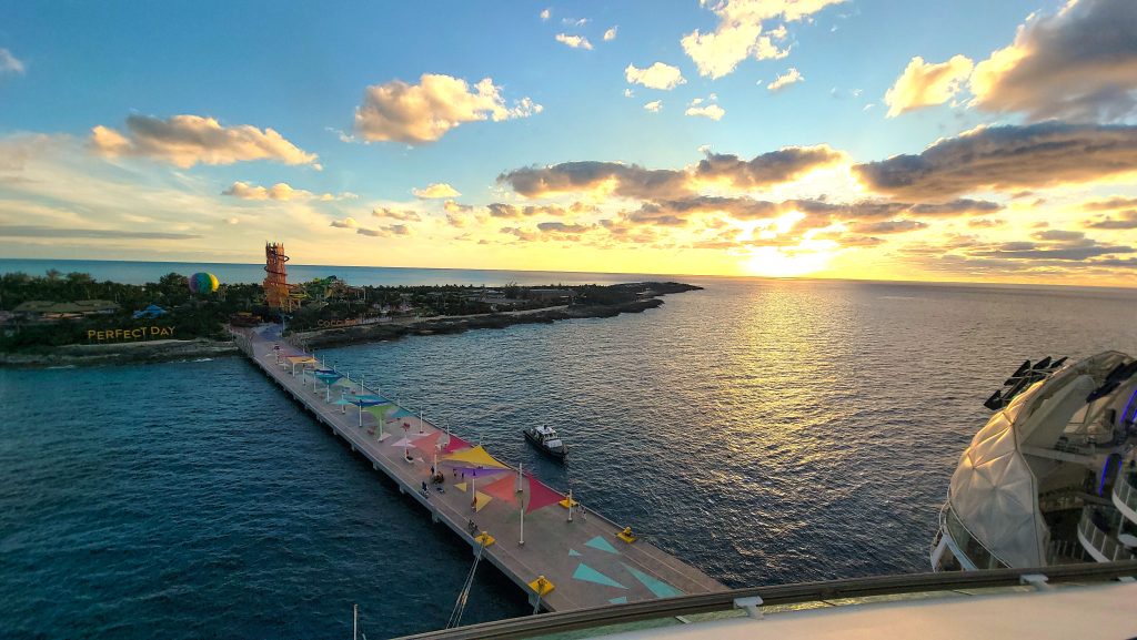 An elevated view from a Royal Caribbean cruise ship looking down at the long arrivals pier with colorful awnings at Perfect Day at CocoCay in the Bahamas during a brilliant golden sunset over the ocean.