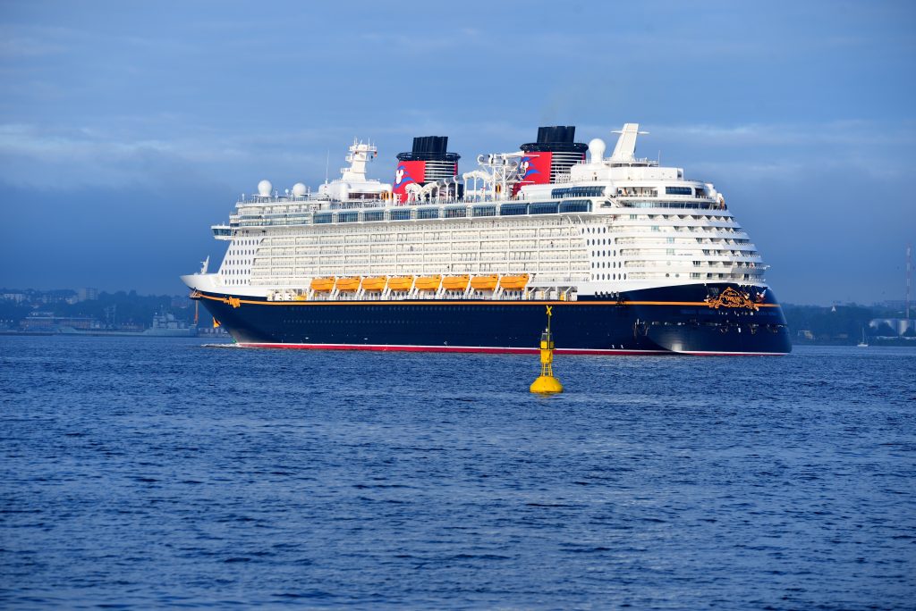A full side profile view of the Disney Dream cruise ship sailing on deep blue water, highlighting its classic black hull, twin red and black funnels with the iconic Mickey Mouse logo, and yellow lifeboats.