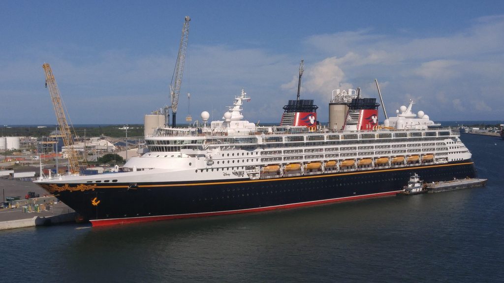 A side view of the Disney Cruise Line ship Disney Wonder docked at a port terminal, featuring its classic dark hull with yellow bow scrollwork and twin red funnels bearing the Mickey Mouse silhouette.