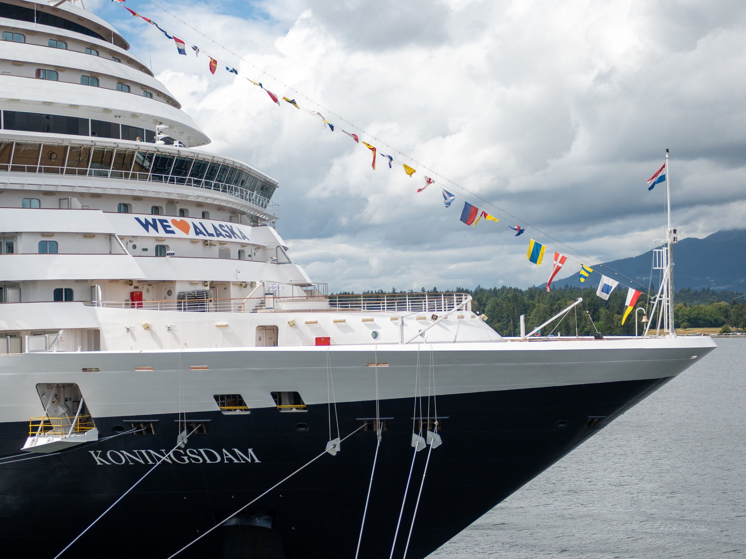 A close-up shot of the bow of the Holland America Line cruise ship MS Koningsdam docked in Alaska, decorated with colorful maritime flags and featuring a prominent "WE ❤️ ALASKA" graphic on its hull.