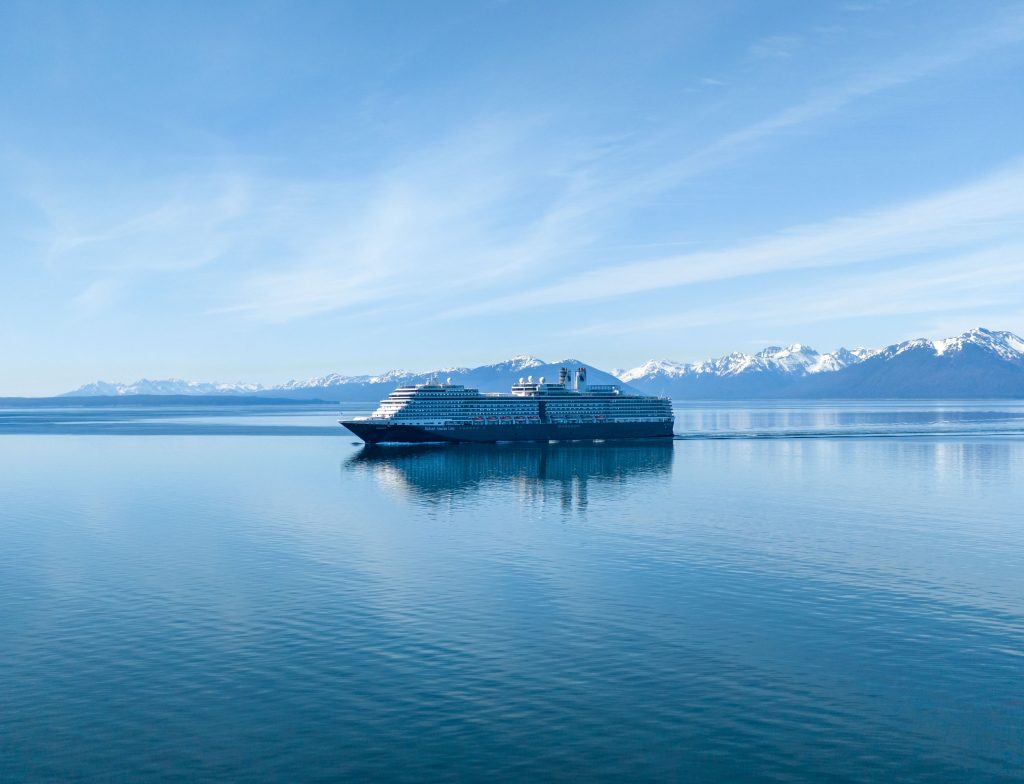 A wide panoramic shot of a dark-hulled Holland America Line cruise ship sailing smoothly through glassy, reflective blue water near Hoonah, Alaska, against a massive backdrop of snow-capped mountains.