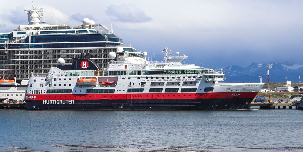 A close-up side view of the HX Hurtigruten Expeditions polar ship, MS Fram, docked in Ushuaia Harbor, Argentina, showcasing its signature red, black, and white hull with destinations like Antarctica, Greenland, and Spitsbergen painted on the side.
