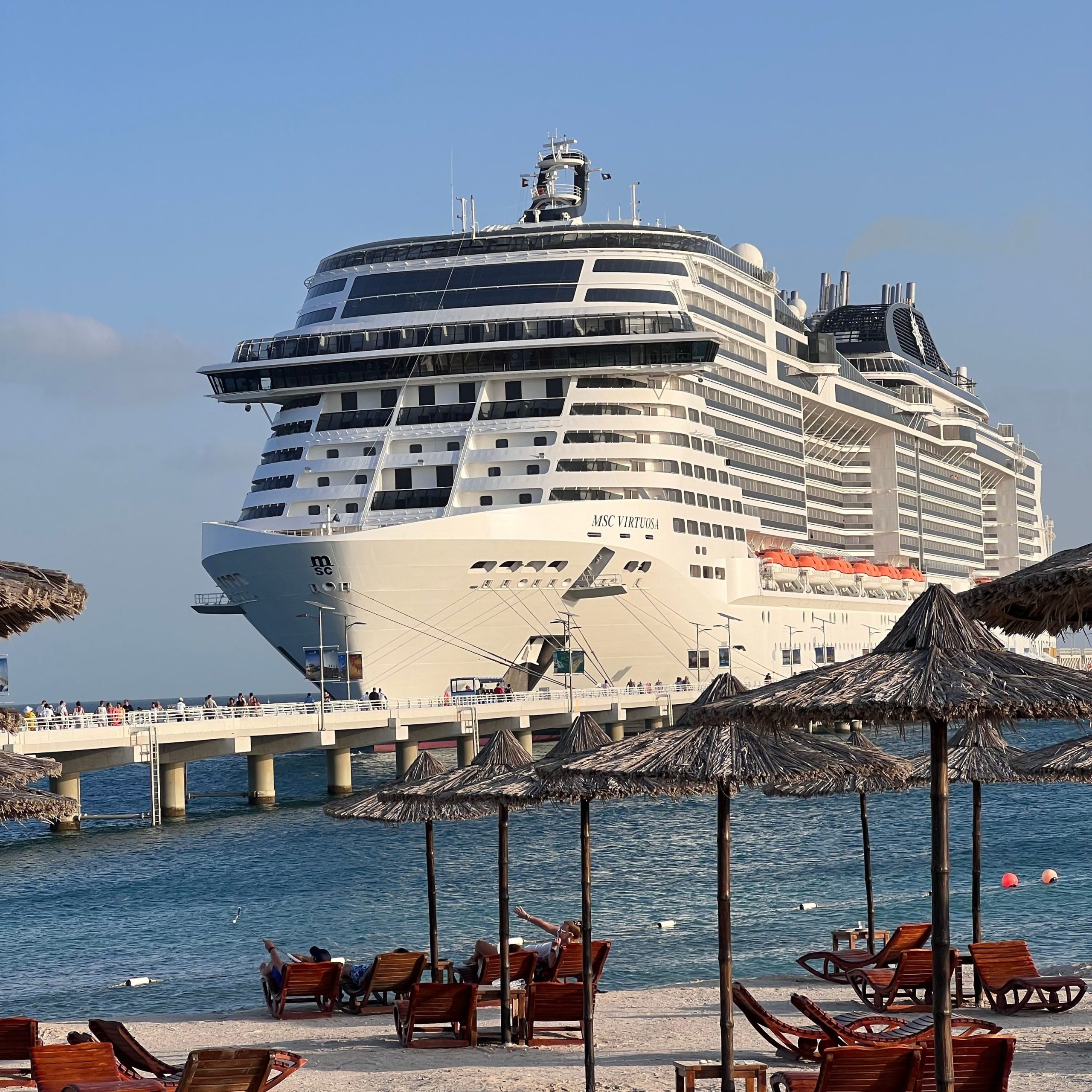 A towering front bow view of the white cruise ship MSC Virtuosa docked at a pier, viewed from a sandy beach lined with wooden lounge chairs and straw-thatched umbrellas under a clear blue sky.