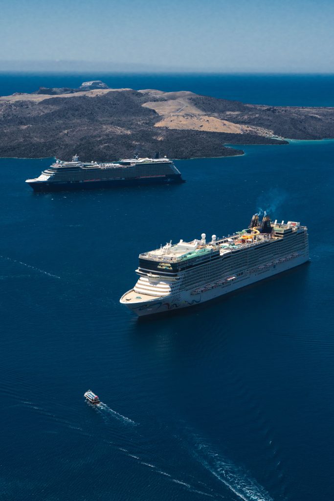 A high aerial view looking down at the Norwegian Epic and Celebrity Equinox cruise ships anchored in the deep blue Aegean Sea off the rugged volcanic coastline of Fira, Greece.