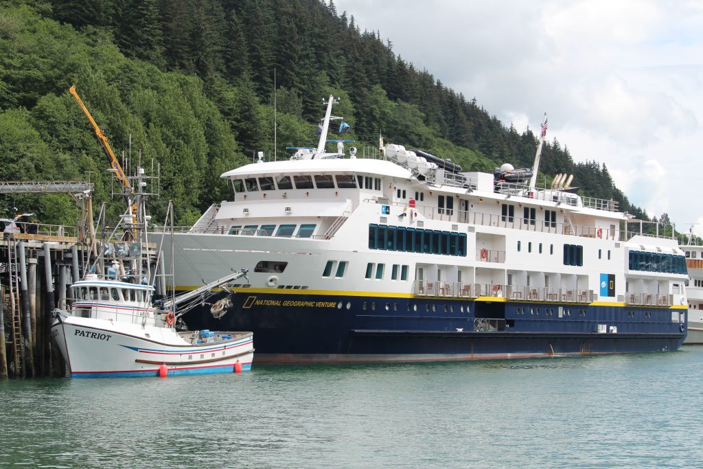 A starboard side view of the white and navy blue expedition ship National Geographic Venture docked at a wooden pier in Skagway, Alaska, with a small white fishing boat named Patriot in the foreground and a steep hill covered in green pine trees in the background.