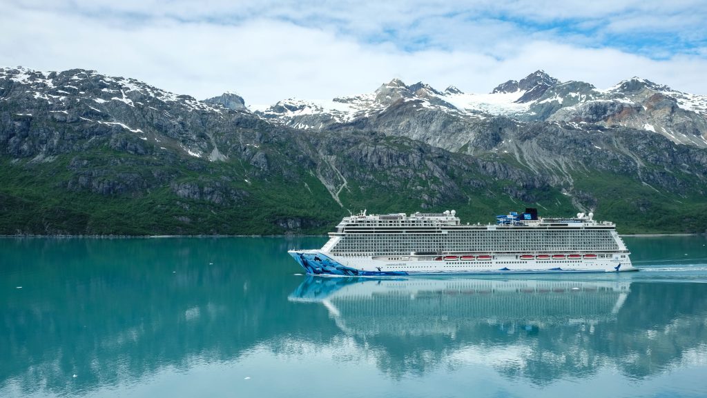The Norwegian Bliss cruise ship sailing through calm, turquoise Alaskan waters with massive snow-capped mountains and green slopes perfectly reflecting in the ocean.