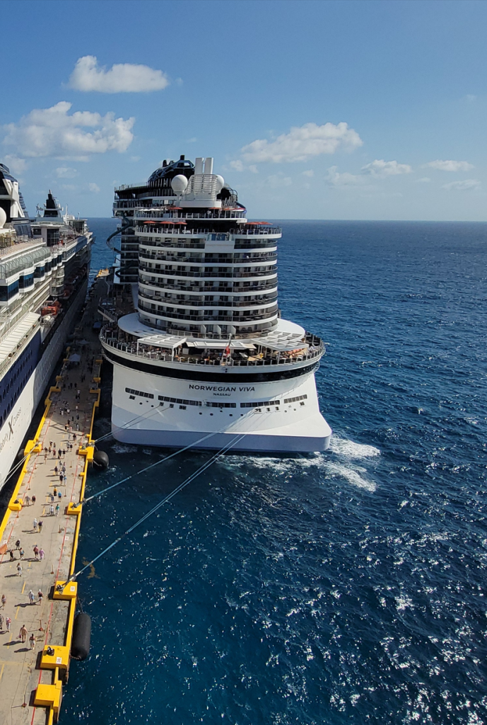 A high-angle overhead view looking down at the unique tiered stern of the Norwegian Cruise Line ship, Norwegian Viva, docked at a long concrete pier with passengers walking alongside the deep blue ocean.
