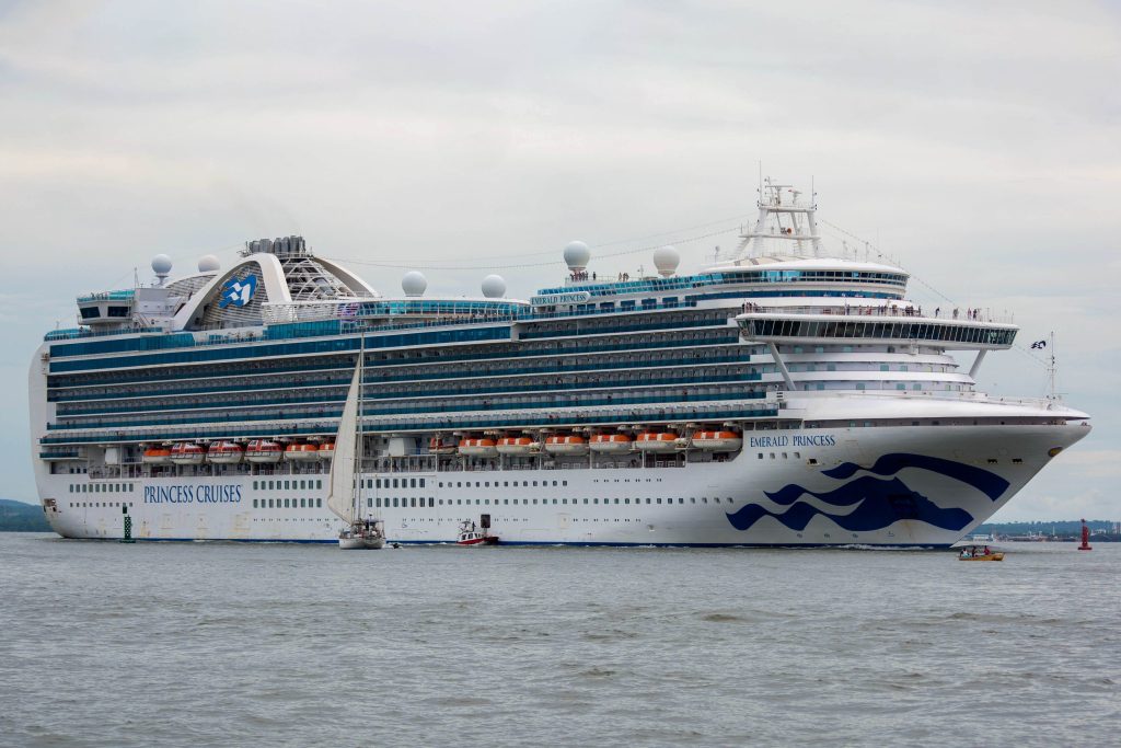 A full port side profile view of the Princess Cruises ship, Emerald Princess, traveling down a shipping channel under a cloudy sky, featuring its iconic blue sea witch logo on the bow with a small white sailboat in the foreground and a tree-lined shore in the distance.