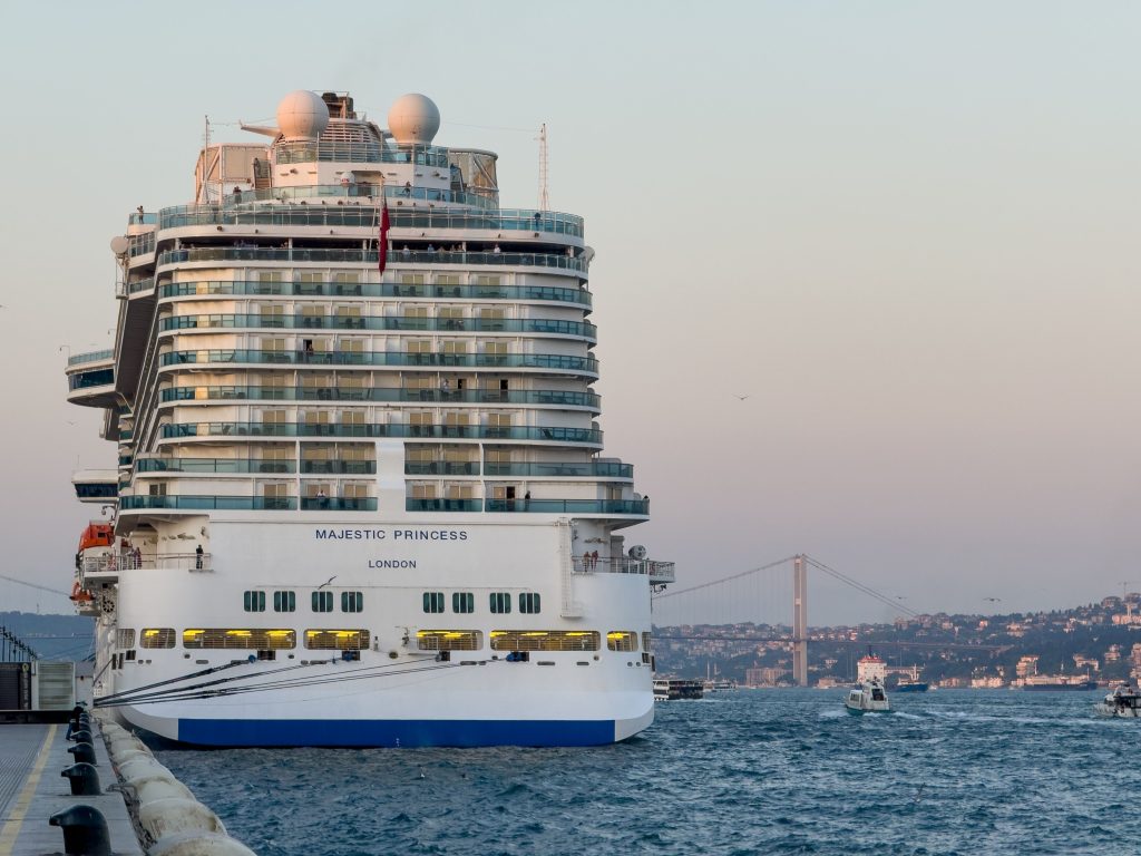 A close-up view of the massive white stern of the Princess Cruises ship, Majestic Princess, docked at a pier in Istanbul, Turkey, with its name clearly visible, while the historic Bosphorus Bridge stretches across the water in the hazy background.