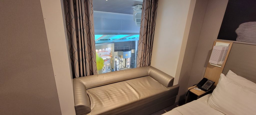 Interior view of a Royal Caribbean promenade stateroom on Harmony of the Seas featuring a small sofa built into a large window alcove that overlooks the brightly lit shops of the Royal Promenade below.