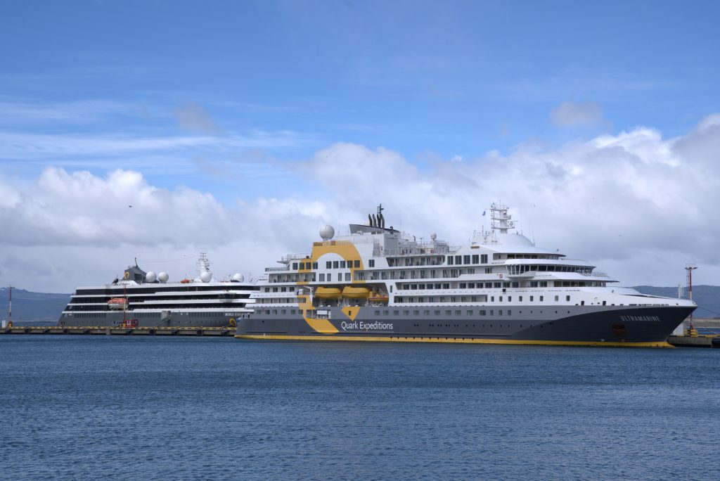A close-up side view of Quark Expeditions' flagship vessel, Ultramarine, docked at a pier in Ushuaia, Argentina under a bright blue sky, showcasing its distinctive grey and yellow hull branding.
