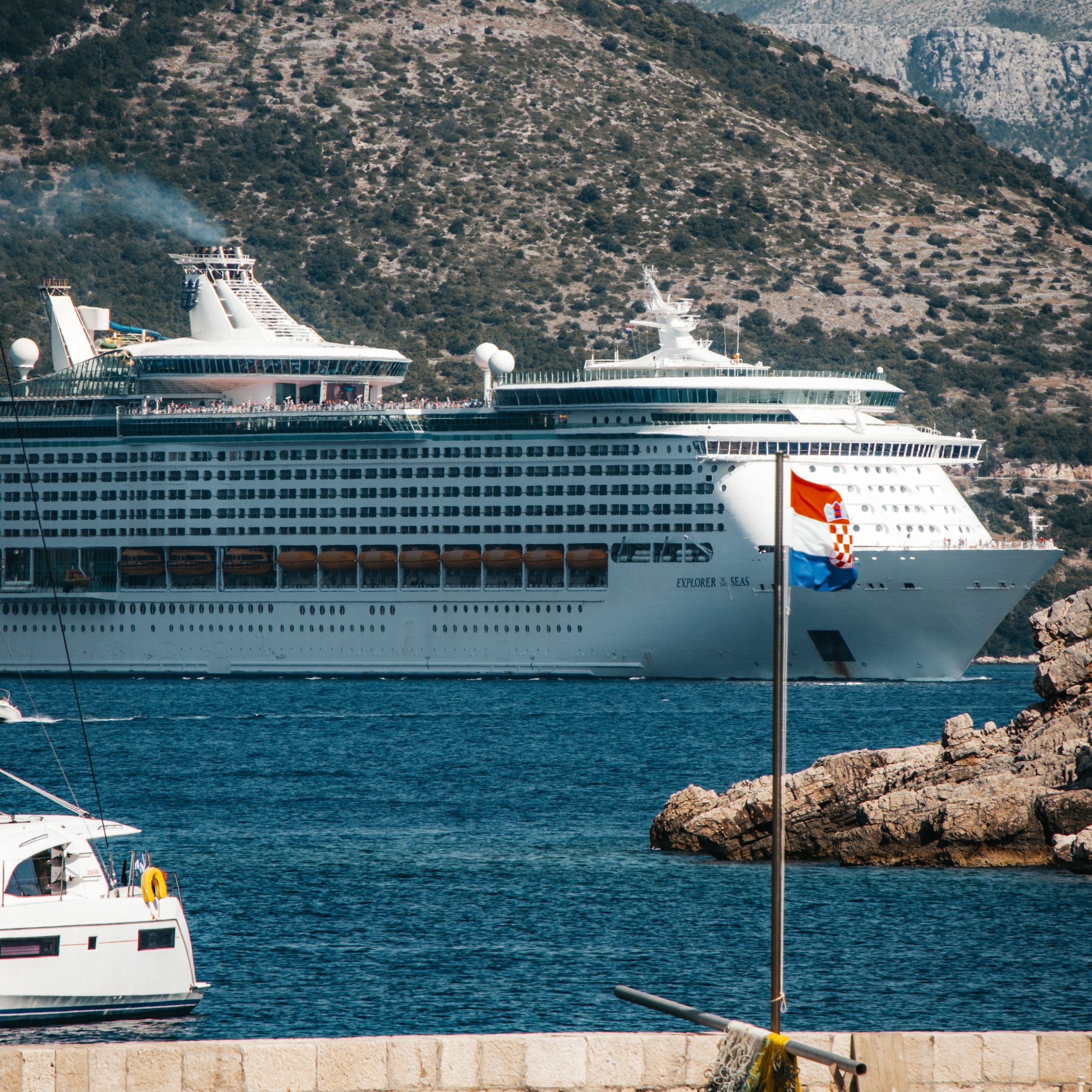 A vertical profile view of the Royal Caribbean cruise ship, Explorer of the Seas, sailing through the deep blue waters of the Adriatic Sea in Dubrovnik, Croatia, with a Croatian flag flying on a stone pier in the foreground and arid mountains in the background.