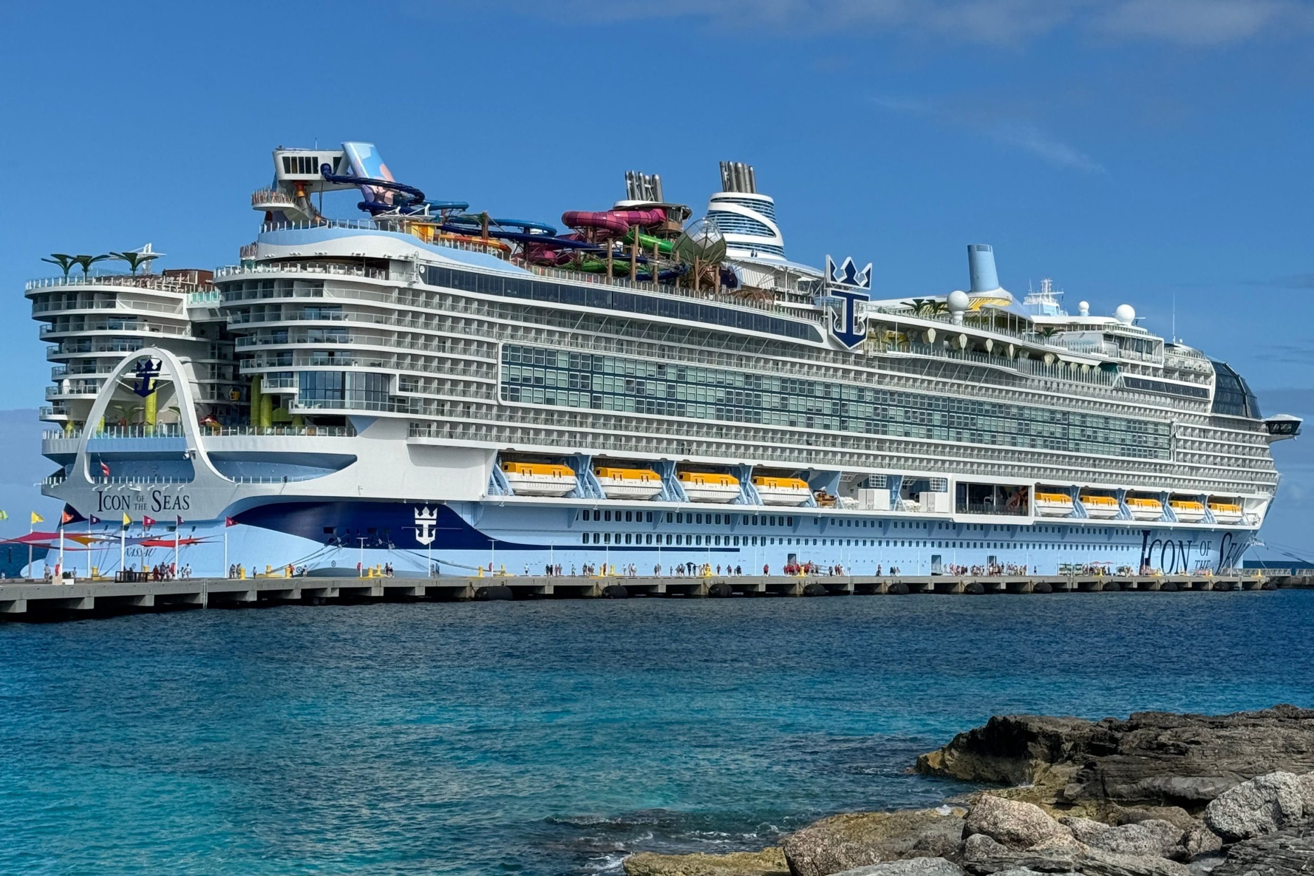 A full side profile view of Royal Caribbean's massive Icon of the Seas docked at a pier, framed by a rocky shoreline and crystal-clear turquoise Caribbean water under a bright blue sky.