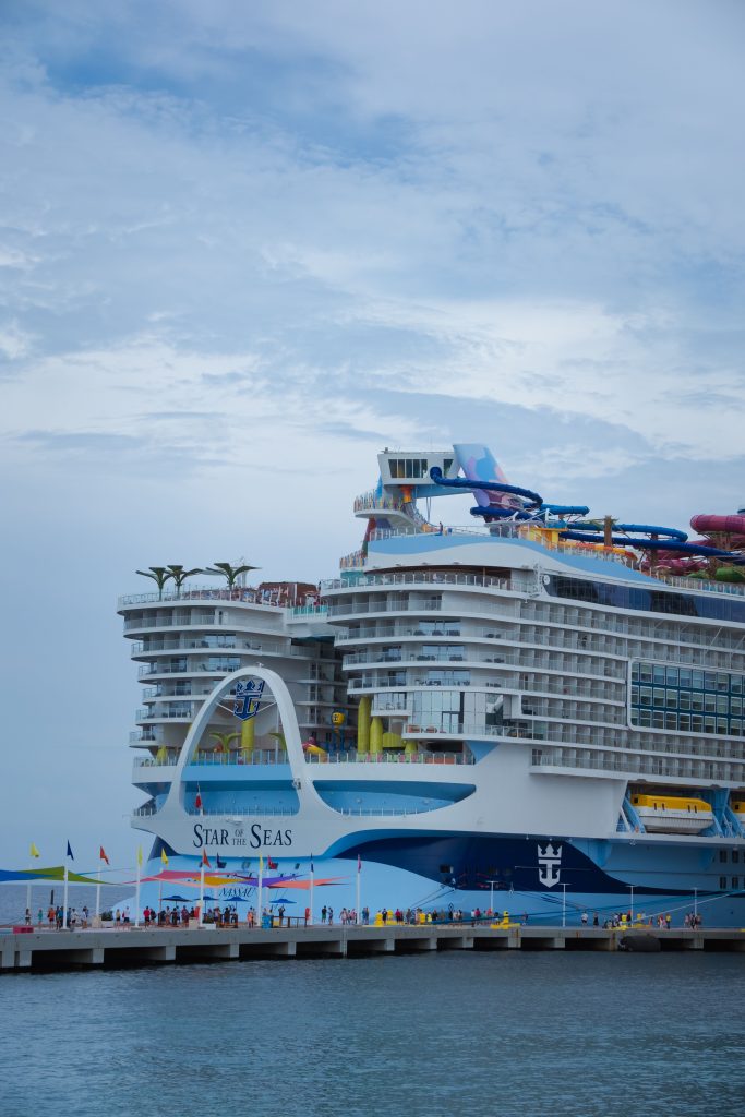 A close-up aft view of the Royal Caribbean cruise ship Star of the Seas docked at a pier in the Bahamas, showcasing its multi-story balcony staterooms, vibrant blue hull, and top-deck waterslides under a cloudy sky.