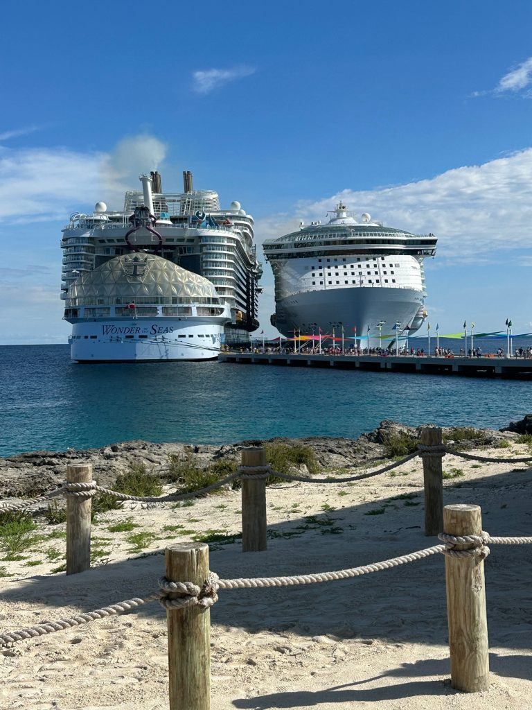 A view from a sandy beach with wooden posts and a rope fence looking out toward the turquoise ocean where Royal Caribbean's Wonder of the Seas is docked side-by-side with a fleet mate at a pier in the Bahamas.