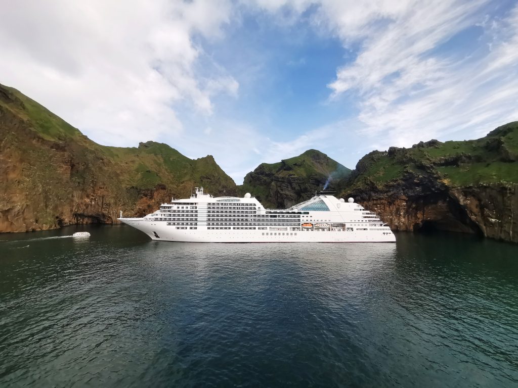 A wide landscape view of the ultra-luxury cruise ship Seabourn Ovation anchored in the calm, dark waters of a dramatic volcanic cove in Vestmannaeyjar, Iceland, surrounded by steep green cliffs under a cloudy blue sky.