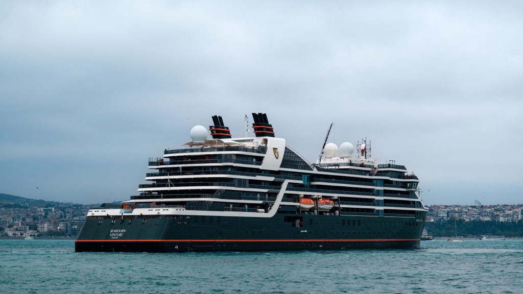 An aft starboard profile view of the ultra-luxury expedition cruise ship Seabourn Venture sailing on calm turquoise water in Istanbul, Turkey, highlighting its dark green and black hull with tiered passenger balconies under a cloudy sky.