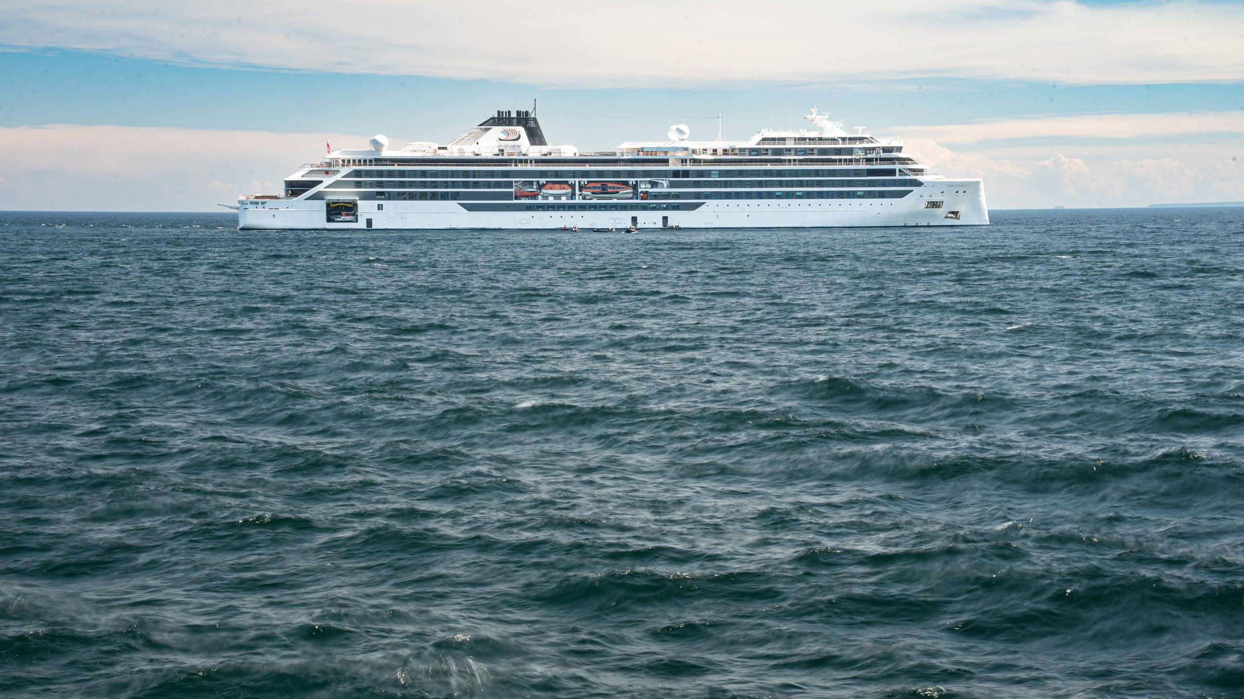 A full side profile view of the sleek white Viking Expeditions ship, Viking Polaris, navigating rolling deep blue ocean waves under a bright, partially cloudy sky.