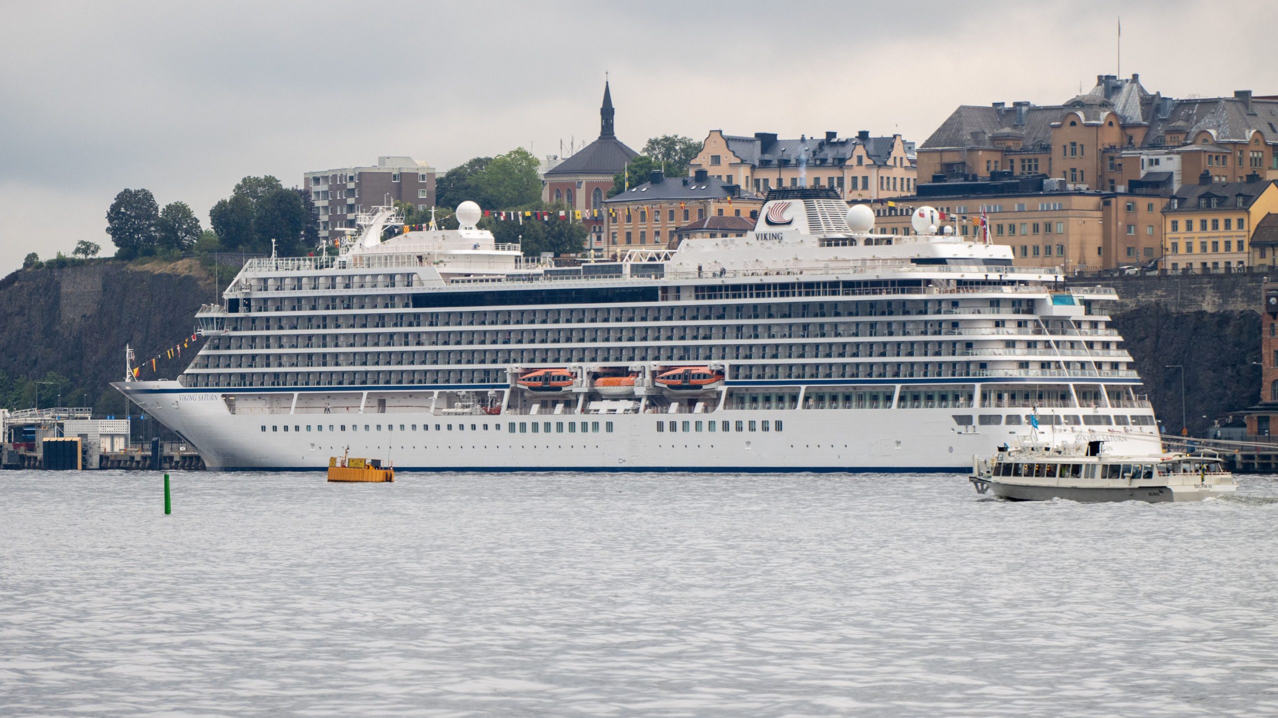 A full starboard side view of the white luxury ocean ship, Viking Saturn, floating in Stockholm Harbor, Sweden under an overcast sky, with a historic European city skyline and a cliffside building complex in the background.