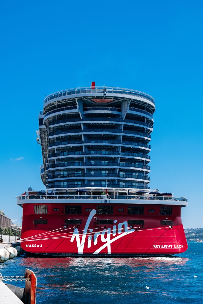 A close-up aft view of Virgin Voyages' Resilient Lady cruise ship docked at Galataport in Istanbul, Türkiye, showcasing its distinct bright red stern emblazoned with the white script Virgin logo under a brilliant blue sky.