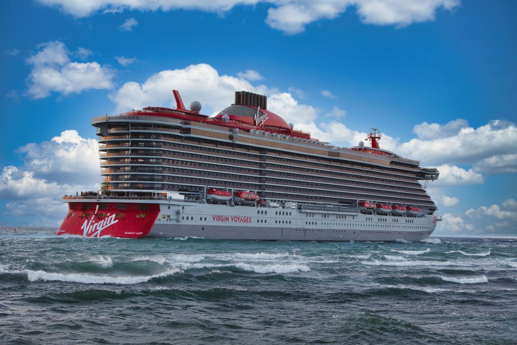 A side-profile shot of the Virgin Voyages cruise ship Scarlet Lady navigating deep blue, choppy ocean waters near Puerto Plata, Dominican Republic, under a beautiful cloudy sky.