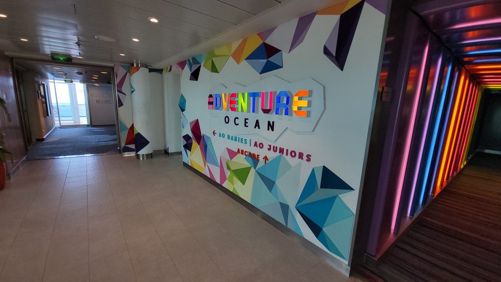 A bright, colorful corridor entrance to the Adventure Ocean youth club on a Royal Caribbean cruise ship, featuring a geometric, multi-colored wall design and glowing rainbow neon lighting leading into the children's activity area.