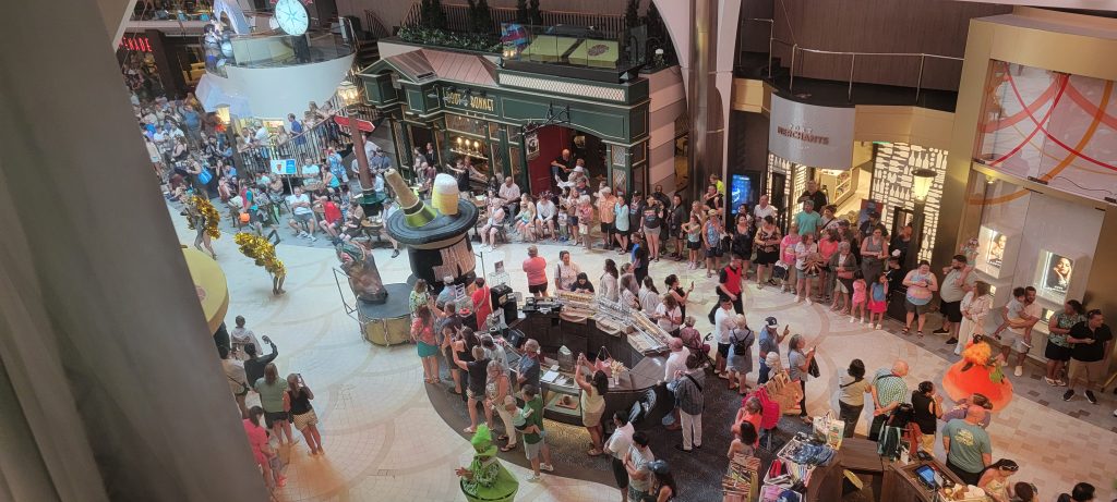 Looking down from a stateroom window at a large, festive crowd gathered on the multi-story Royal Promenade of Harmony of the Seas, featuring passengers watching a celebration near the onboard bars, shops, and a giant decorative top-hat display.