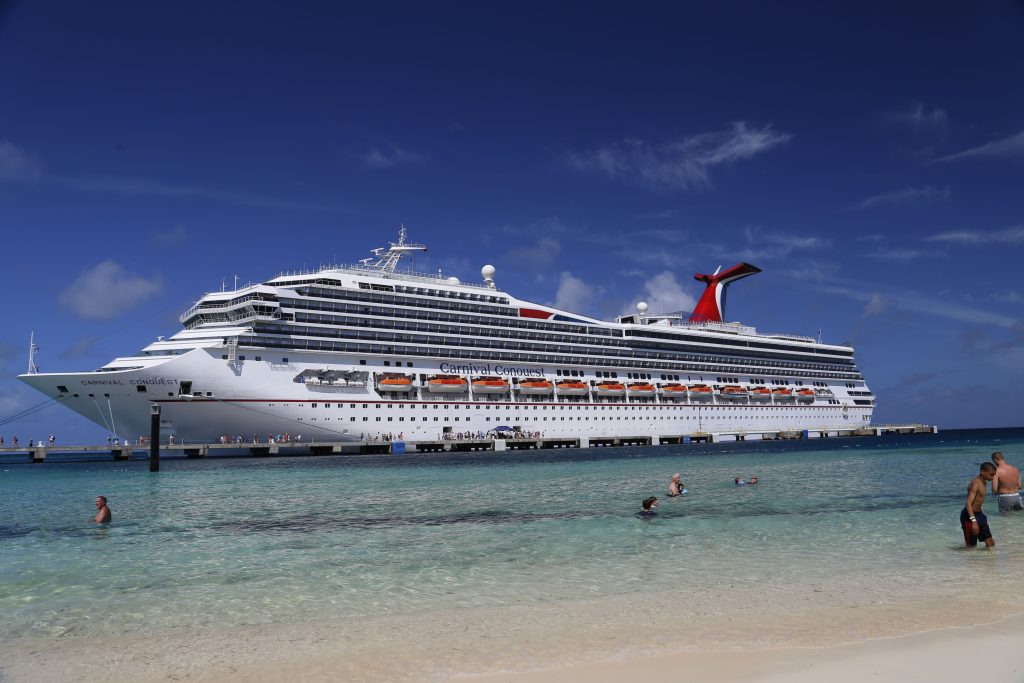 A side profile view of the Carnival Conquest cruise ship docked at a long pier in the Bahamas, seen from a sandy beach with people swimming in the clear, shallow turquoise water under a blue sky.