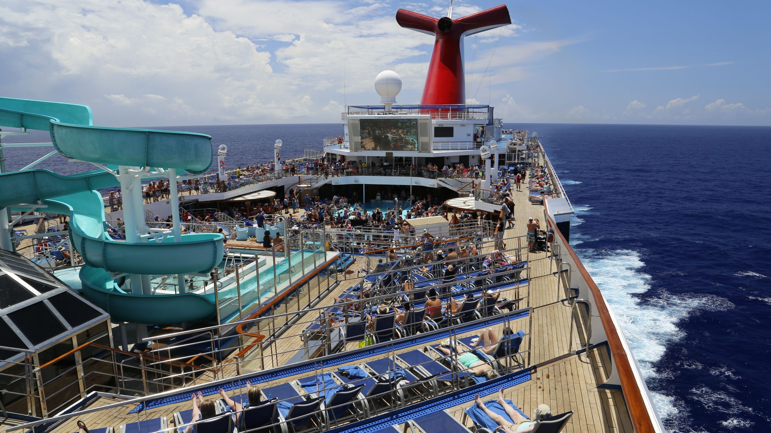 Looking down on the sunny outdoor top deck of a Carnival cruise ship at sea, featuring a large teal waterslide, rows of blue lounge chairs with passengers sunbathing, a crowded mid-ship pool area, and the iconic red and blue whale-tail funnel.