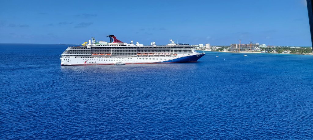 A beautiful, full side profile view of the Carnival Spirit cruise ship anchored in the deep blue waters of the Cayman Islands under a clear sky.