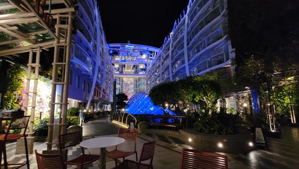 A vibrant night view looking through the lush, plant-filled Central Park neighborhood on Royal Caribbean's Harmony of the Seas, featuring a glowing blue glass structure and warm lighting from surrounding balcony staterooms.