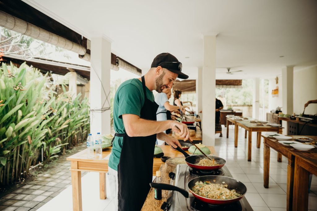 An enthusiastic participant in an outdoor cooking class, wearing a black apron and stirring food in a pan over a gas stove, illustrating the hands-on nature of local culinary experiences.