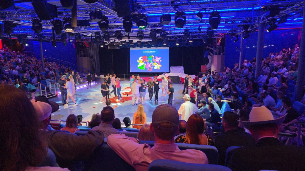 Looking down at the brightly lit Studio B ice rink arena turned into a game show floor on a Royal Caribbean ship, featuring contestants gathered on stage, a large digital screen displaying the colorful Crazy Quest logo, and packed stadium seating filled with a spectator crowd.