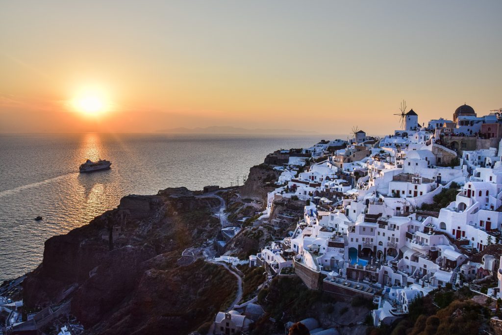 A breathtaking sunset view looking down at the white-washed cliffside buildings and windmills of Oia in Santorini, Greece, with a cruise ship sailing across the golden, glowing waters of the Aegean Sea.