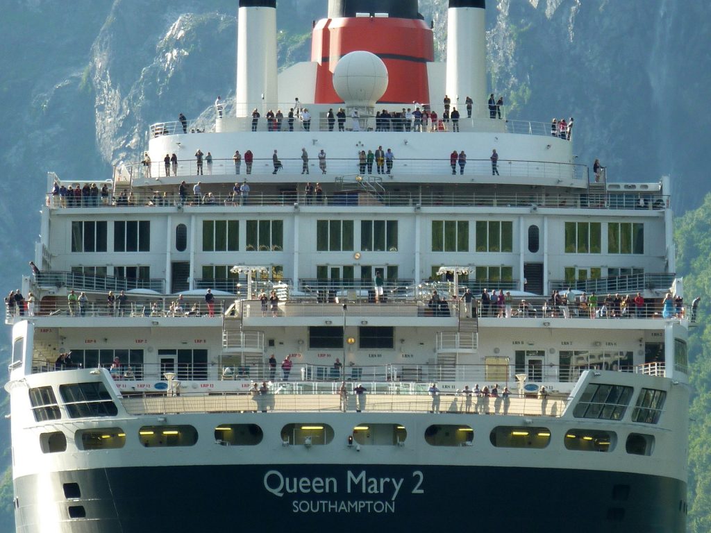 A towering, close-up view looking up at the multi-tiered decks of Cunard's Queen Mary 2 ocean liner, showing passengers standing along the railings under the signature red and black funnel with the ship's name clearly visible on the superstructure.