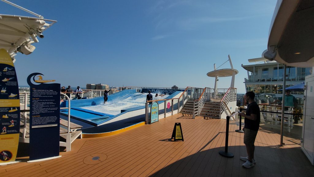 A bright afternoon view of the FlowRider surfing simulator on the wooden sports deck of a Royal Caribbean mega-ship, featuring a passenger riding the artificial wave under a clear blue sky.