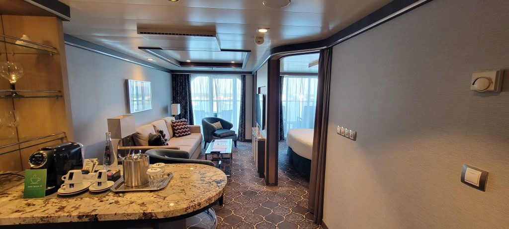 A spacious, luxury cruise ship suite featuring a granite-topped bar with an espresso machine, a separate living room area with a sofa, and a doorway leading into a private bedroom suite.