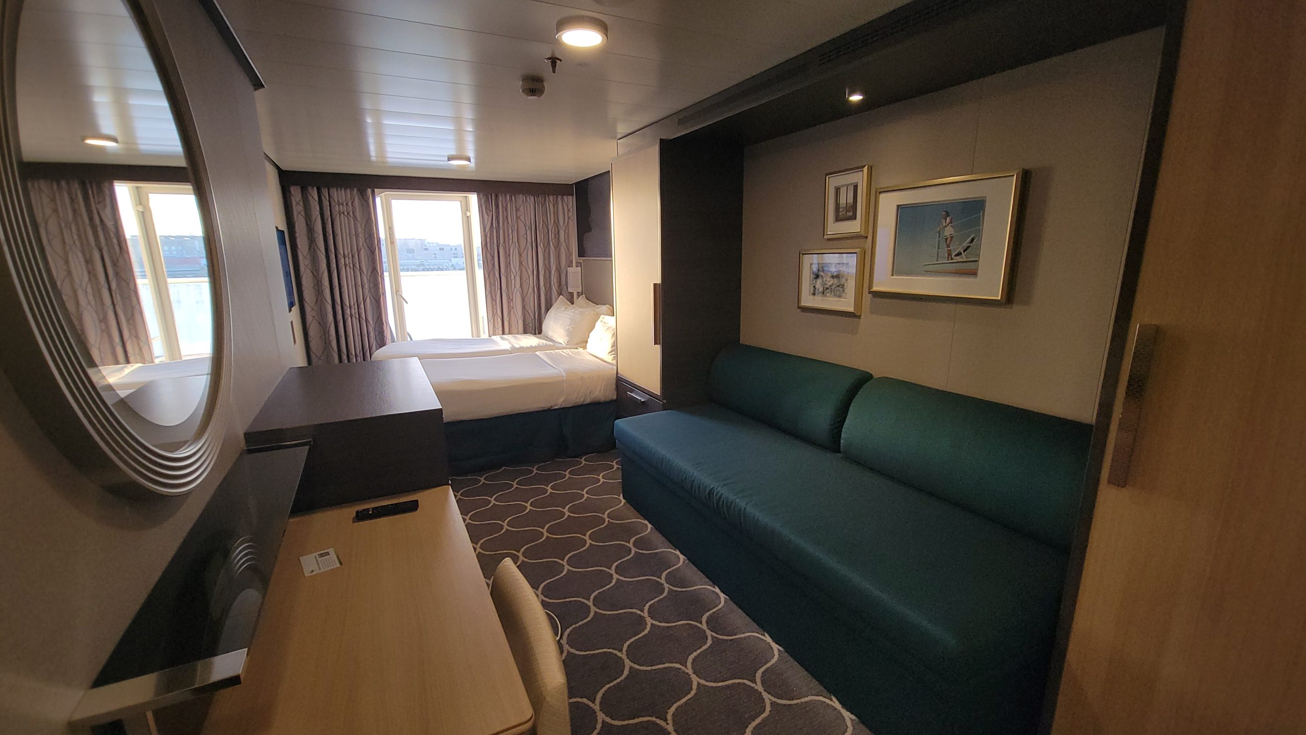 An interior view of a standard Oceanview Balcony stateroom cabin onboard Harmony of the Seas, showing the vanity desk, mirror, teal sofa, and twin beds.