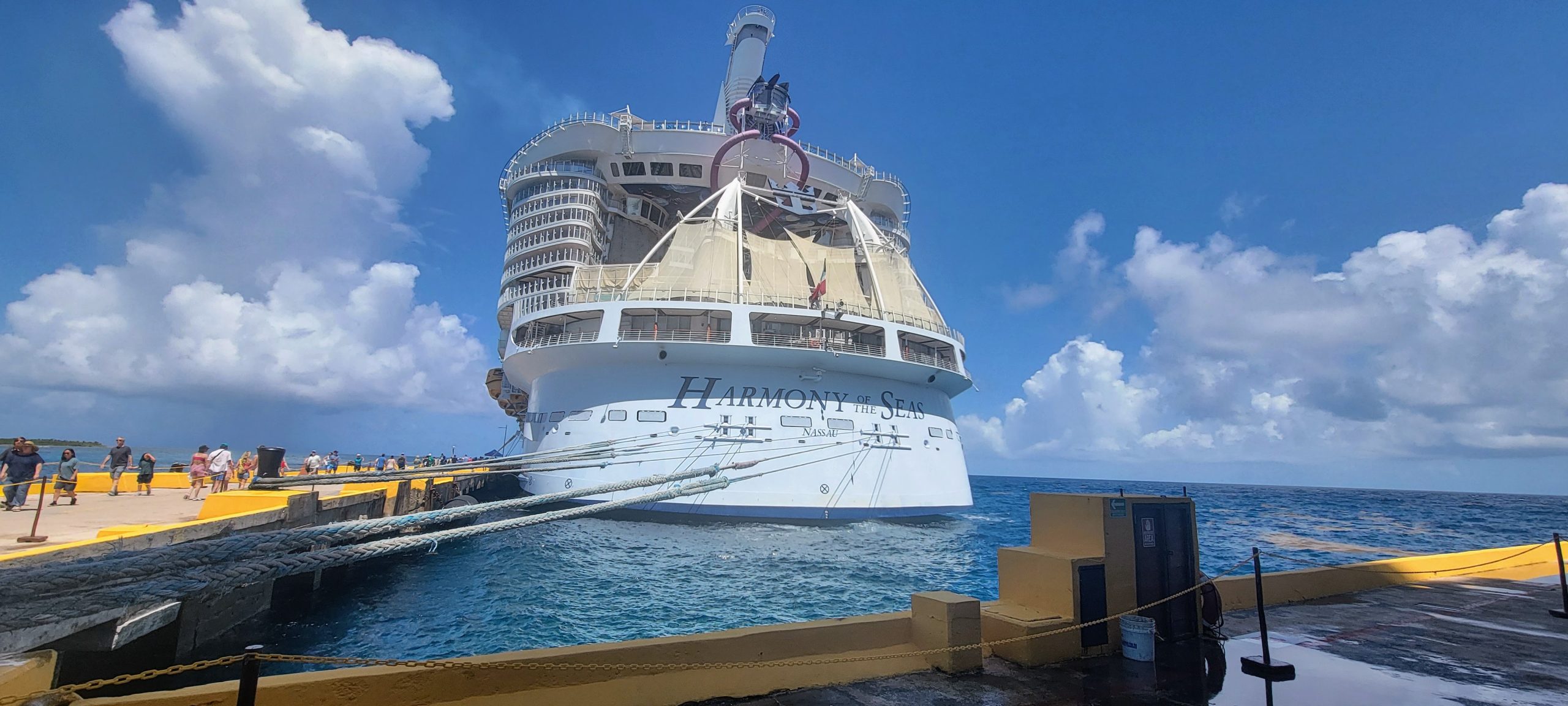 A full aft view of Royal Caribbean's Harmony of the Seas docked at the cruise port pier in Costa Maya, showcasing the ship's massive stern and upper deck structures under a blue sky.