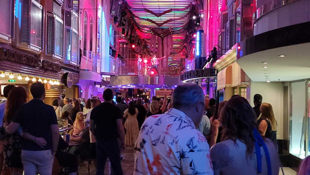 A lively, crowded evening dance party filled with guests on the multi-story Royal Promenade inside Royal Caribbean's Liberty of the Seas, illuminated by vibrant pink and purple venue lighting.