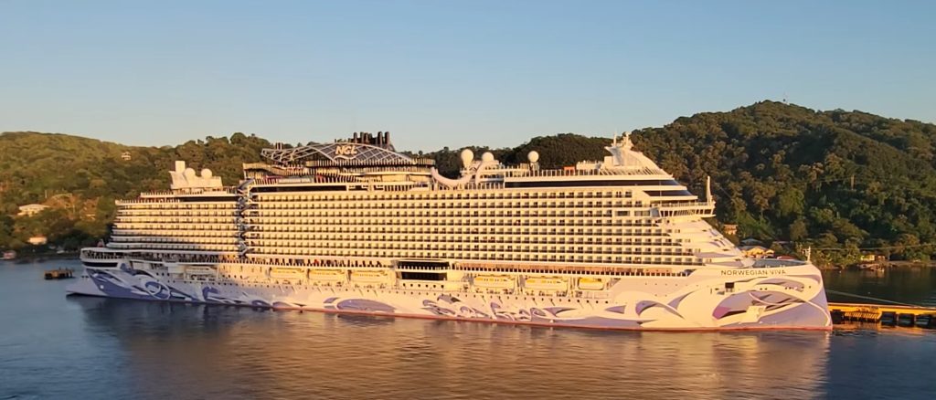 A full side-profile view of the Norwegian Viva cruise ship at sunrise in Roatan, Honduras, showing the morning light illuminating its colorful hull art, rows of balcony cabins, and lush green hills in the background.