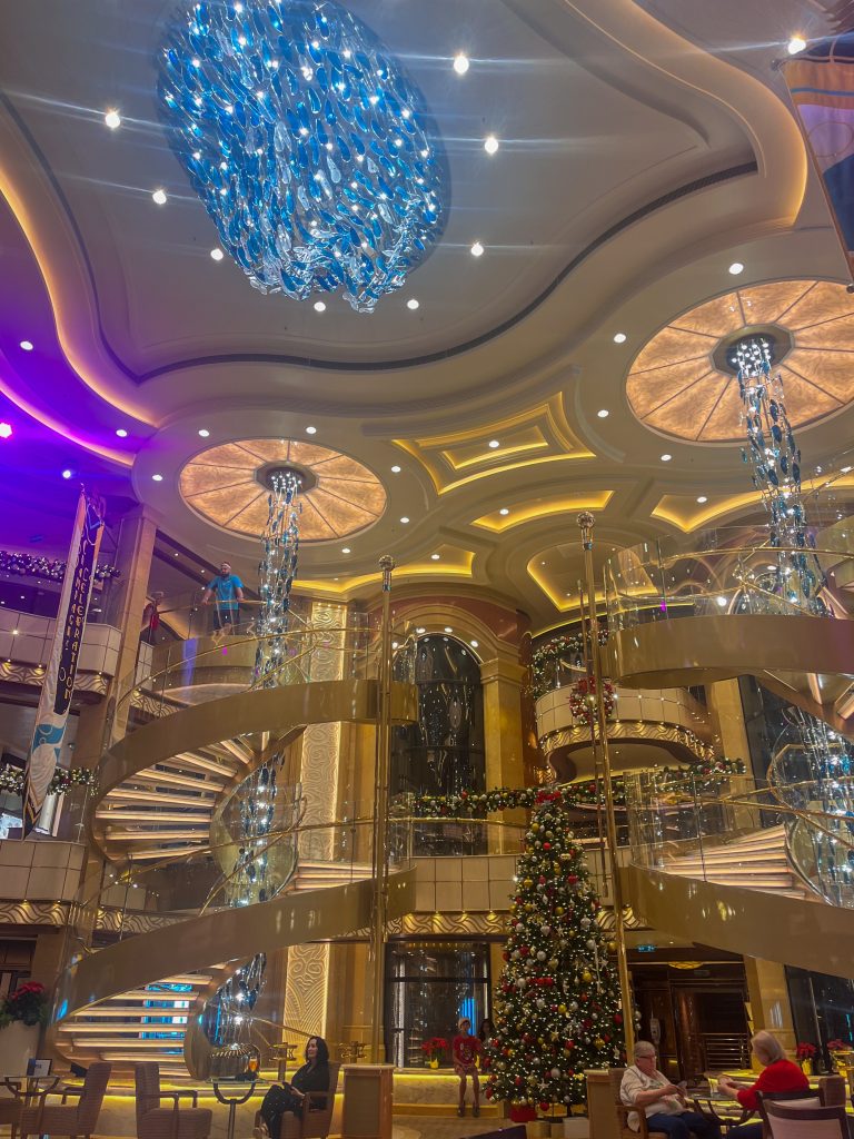 A towering view of the elegant, multi-story Piazza atrium inside a Princess cruise ship, featuring grand golden spiral staircases, festive Christmas trees with warm lights, and a massive, vibrant blue glass chandelier suspended from the polished ceiling.