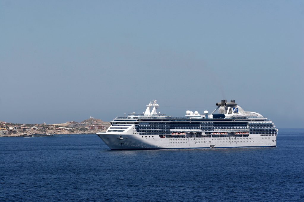 A side profile view of the mid-sized cruise ship Island Princess sailing through deep blue ocean waters near a rocky, mountainous coastline under a clear sky.